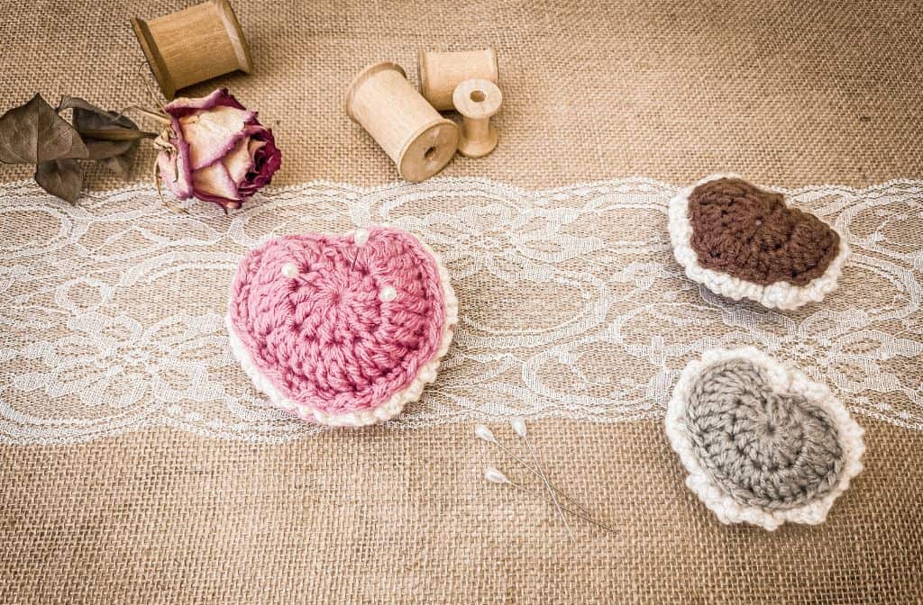 Rustic flat lay of three crochet puffed heart keepsakes in pink, brown, and sage green, displayed on lace over burlap. The pink heart is used as a pin cushion with pearl-topped pins. Surrounded by wooden spools, a dried rose, and decorative pins.