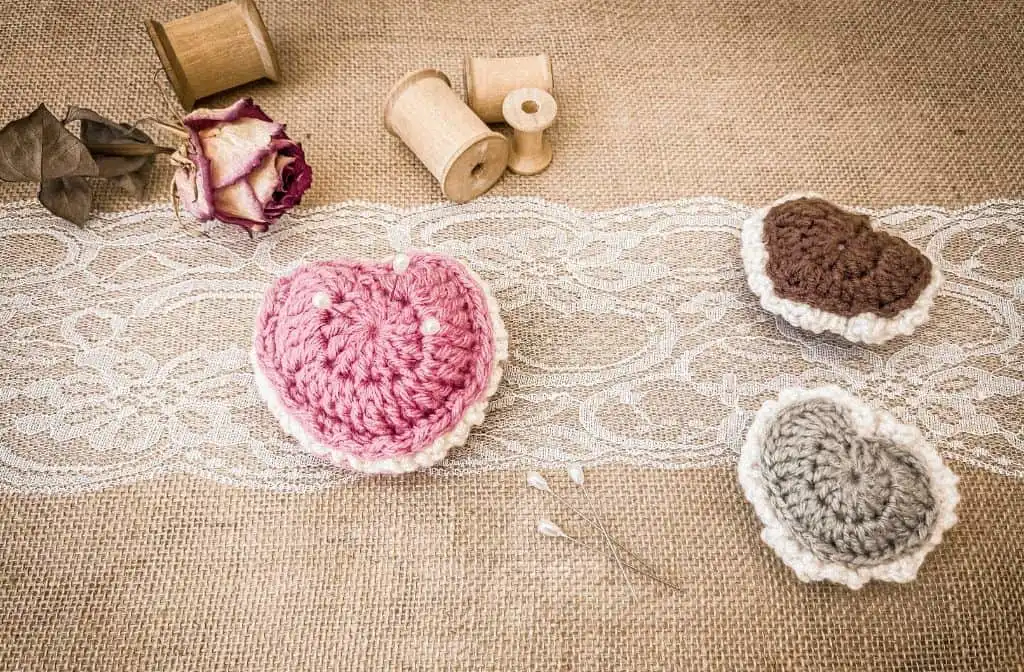 Rustic flat lay of three crochet puffed heart keepsakes in pink, brown, and sage green, displayed on lace over burlap. The pink heart is used as a pin cushion with pearl-topped pins. Surrounded by wooden spools, a dried rose, and decorative pins.