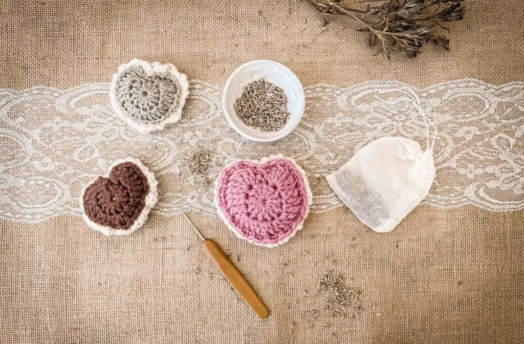 Flat lay of three crochet puffed heart sachets in pink, brown, and sage green, surrounded by dried lavender, a small crochet hook, a lace strip, and a muslin bag on burlap fabric. Rustic and vintage aesthetic
