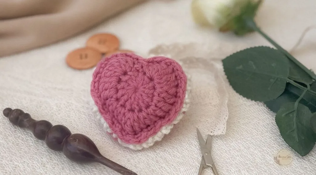 Flat lay of a pink and white crochet puffed heart keepsake on a lace surface, surrounded by vintage-inspired crafting tools, including small scissors, a wooden crochet hook, and large wooden buttons.