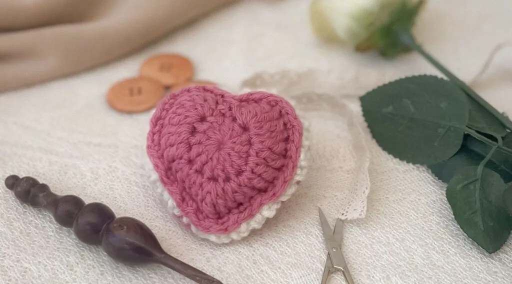 Flat lay of a pink and white crochet puffed heart keepsake on a lace surface, surrounded by vintage-inspired crafting tools, including small scissors, a wooden crochet hook, and large wooden buttons.