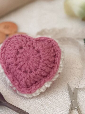 Flat lay of a pink and white crochet puffed heart keepsake on a lace surface, surrounded by vintage-inspired crafting tools, including small scissors, a wooden crochet hook, and large wooden buttons.