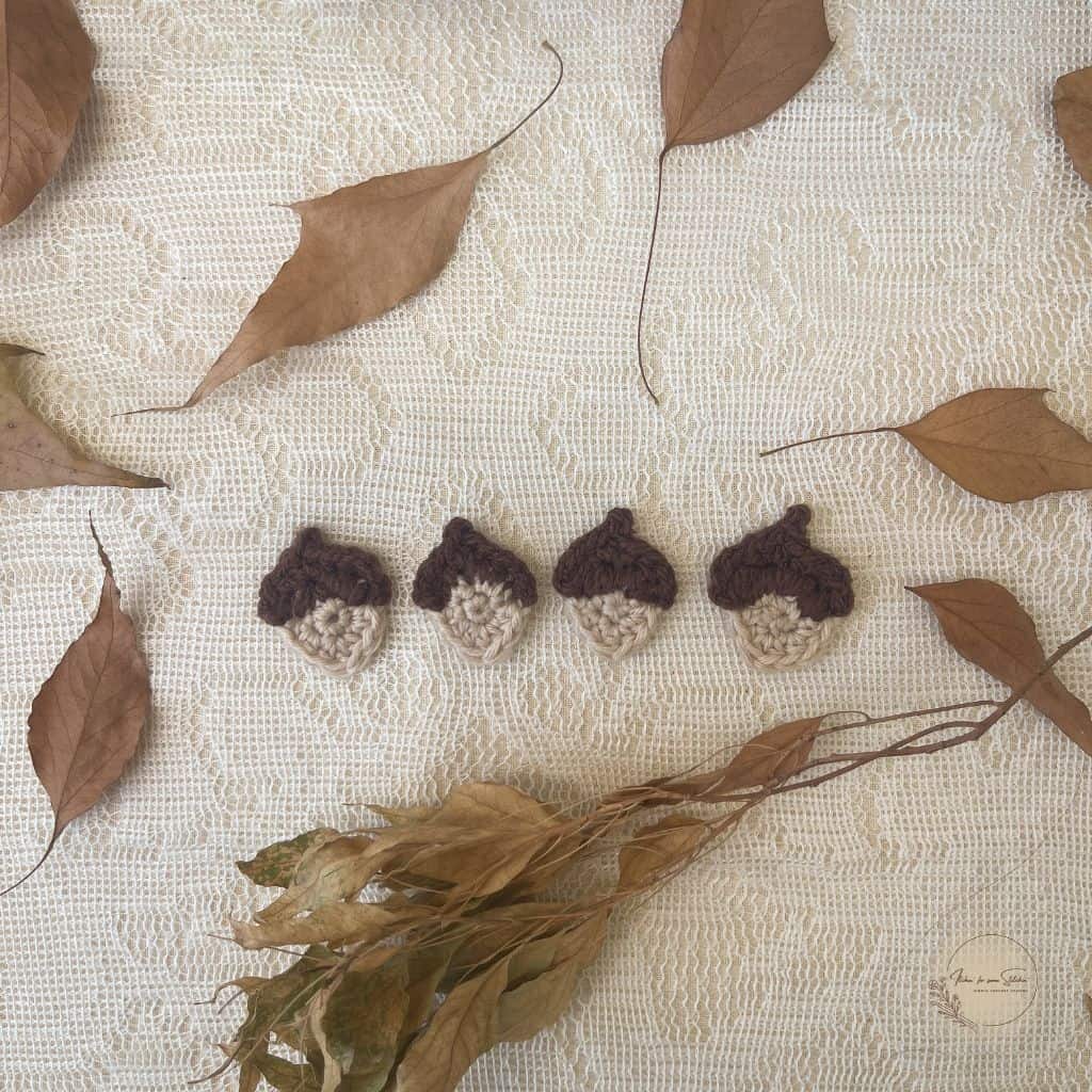Multiple crochet acorn appliqués in brown and beige yarn displayed on a lace background with dried leaves.