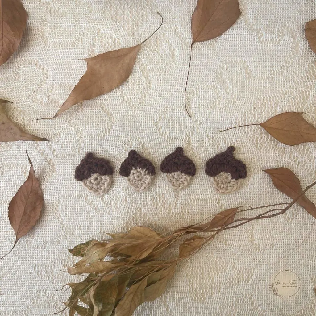 Multiple crochet acorn appliqués in brown and beige yarn displayed on a lace background with dried leaves.