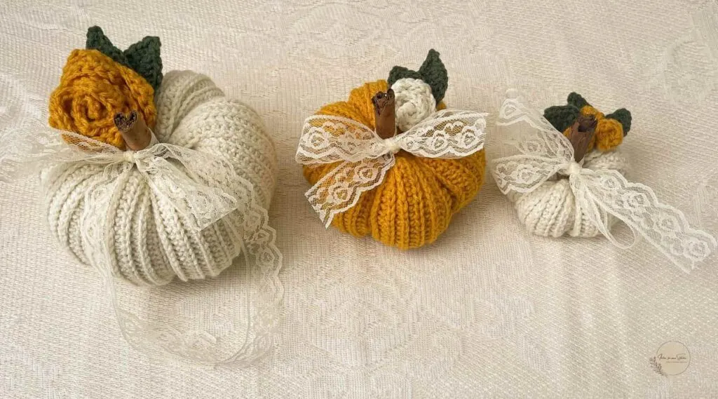 Three ribbed crochet pumpkins in white, mustard, and green with lace bows and cinnamon stick stems displayed on a wooden table — cozy handmade fall décor made with beginner-friendly crochet pattern.