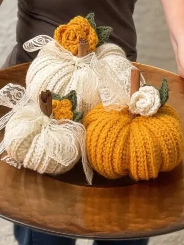 Three ribbed crochet pumpkins in white, mustard, and green with lace bows and cinnamon stick stems displayed on a wooden table — cozy handmade fall décor made with beginner-friendly crochet pattern.