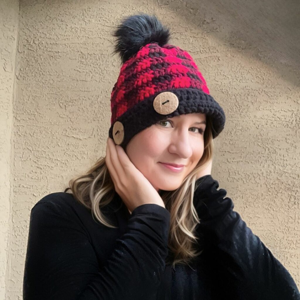 Woman modeling a buffalo plaid crochet hat pattern featuring red, burgundy, and black plaid colorwork with a faux fur pom pom.