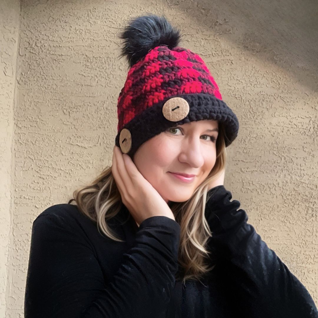 Woman modeling a buffalo plaid crochet hat pattern featuring red, burgundy, and black plaid colorwork with a faux fur pom pom.