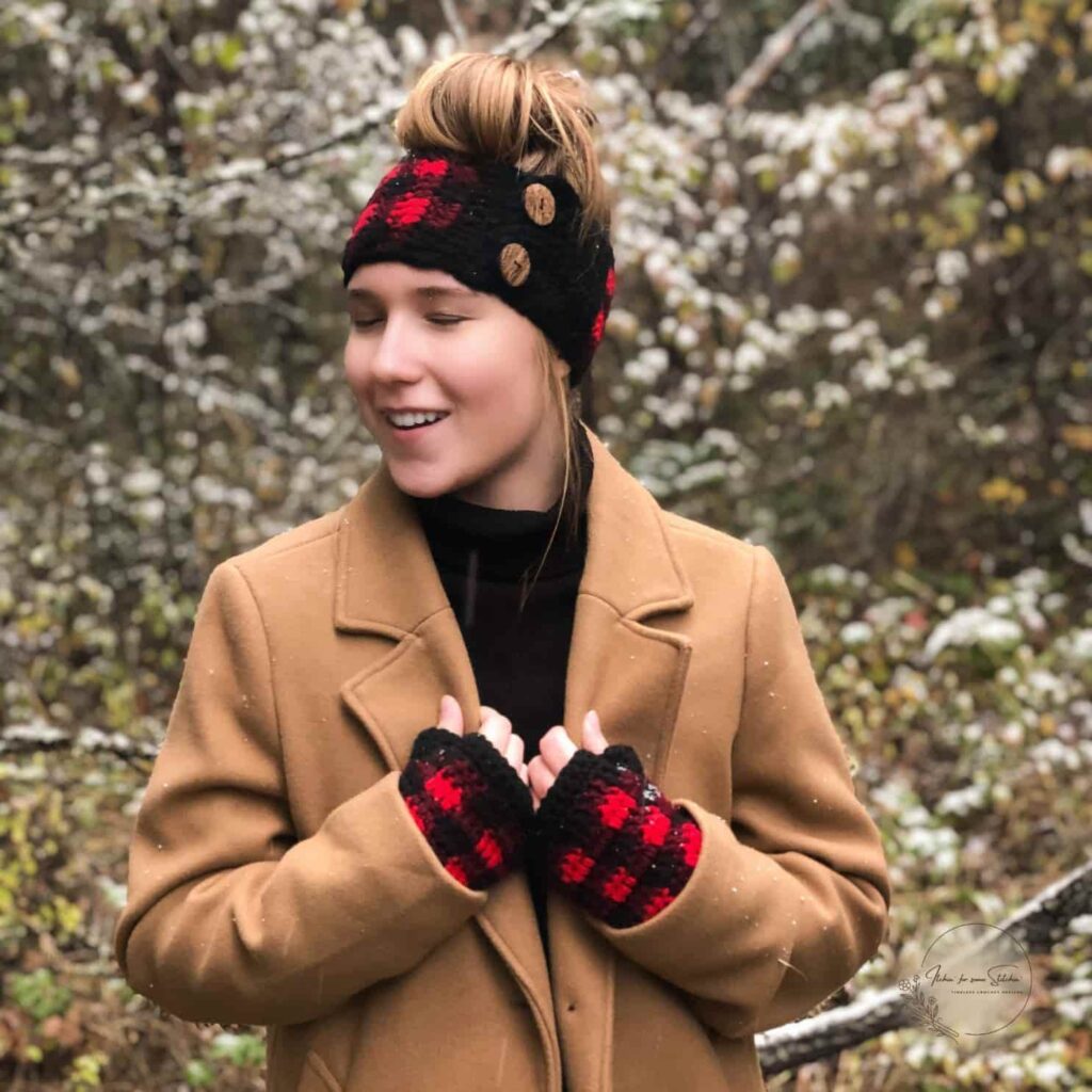 woman wearing a buffalo plaid ear warmer and matching fingerless gloves in red and black while standing outdoors in a camel coat.