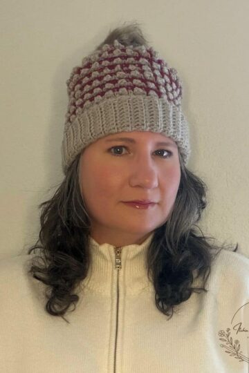 Woman wearing the Everley Beanie, a burgundy and oatmeal crochet hat with raised Mini Berry stitches, a folded ribbed brim, and a faux-fur pom-pom.
