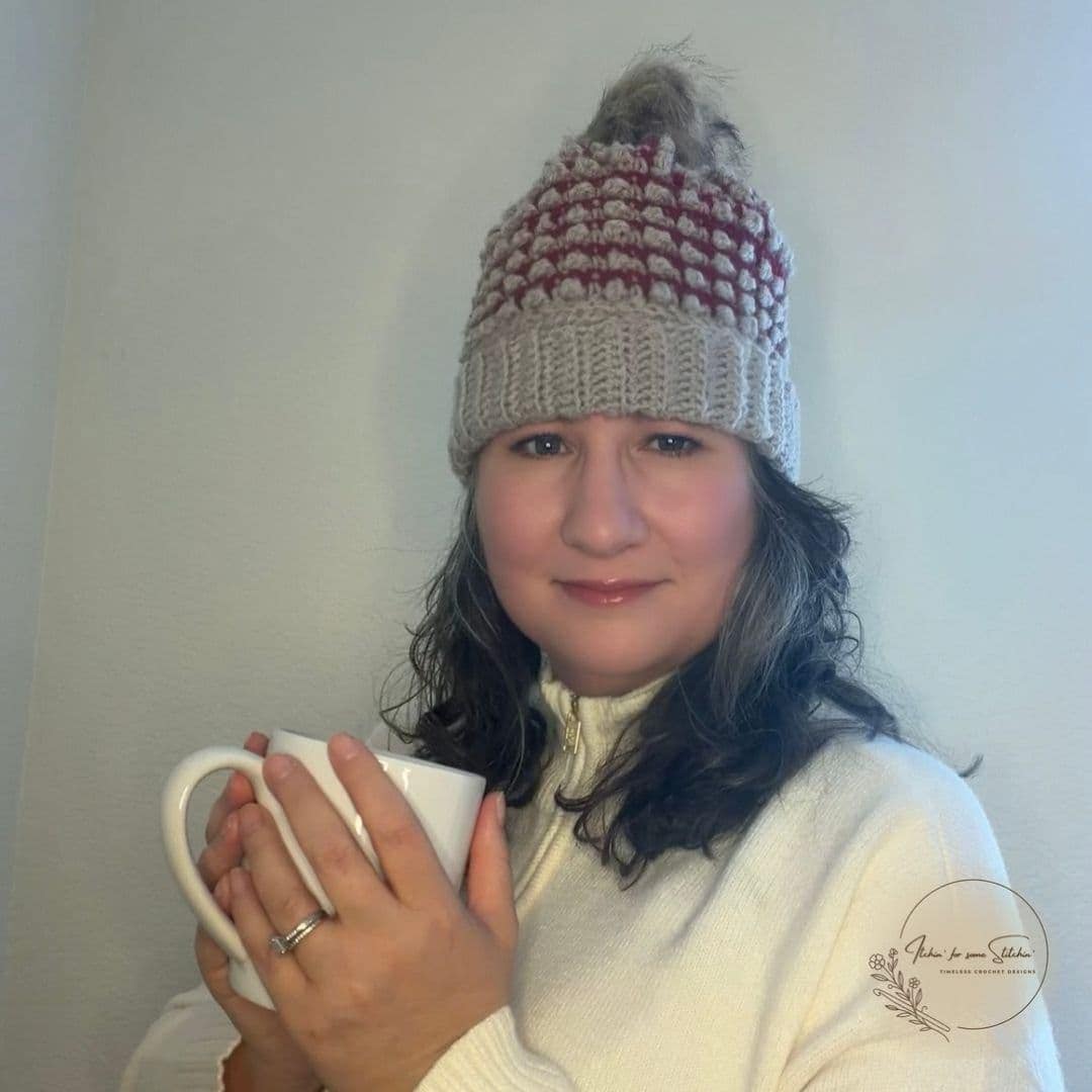 Woman holding a coffee mug while wearing the Everley Beanie, a burgundy and oatmeal textured crochet hat with a folded ribbed brim and faux-fur pom-pom.